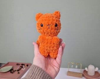 May include: A small orange crocheted cat toy with black eyes and whiskers. The cat is being held in a hand wearing a brown and black striped sweater.