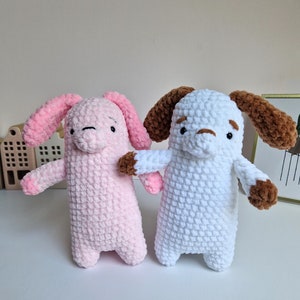 Op de afbeelding: Twee handgemaakte gehaakte knuffels. Eén is roze met roze oren en bruine accenten, de andere is wit met bruine oren en accenten. Beide hebben kleine zwarte ogen en staan rechtop.