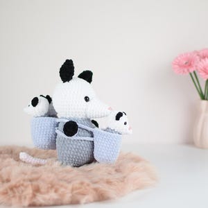 Puede incluir: Un opossum de crochet con dos opossums más pequeños en su bolsa. El opossum es blanco con orejas y ojos negros, y los opossums más pequeños son blancos con ojos negros. El opossum está sentado en una superficie esponjosa de color marrón claro.