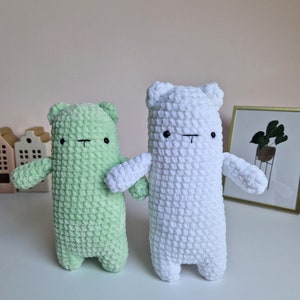 Op de afbeelding: Twee gehaakte knuffels, een groene en een witte beer, met eenvoudige zwarte geborduurde ogen en monden. De beren staan op een wit oppervlak.