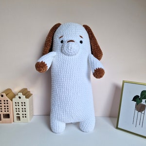 Op de afbeelding: Witte gehaakte knuffel in de vorm van een hond met bruine oren en een bruine neus. Het speelgoed staat op een wit oppervlak.