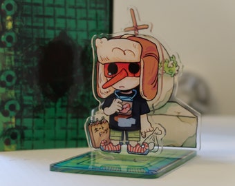 Può includere: Una figura in acrilico trasparente di un personaggio dei cartoni animati con cappello, maschera rossa e un hot dog. La figura è su una base verde e blu. Sullo sfondo c'è un libro verde e nero a motivi.