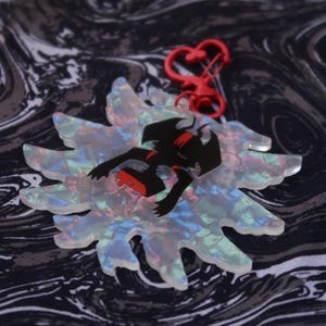 May include: A clear, iridescent acrylic charm in a flower shape with a black figure and red accents. The charm is attached to a red heart-shaped clasp. The background is a black and white swirled pattern.