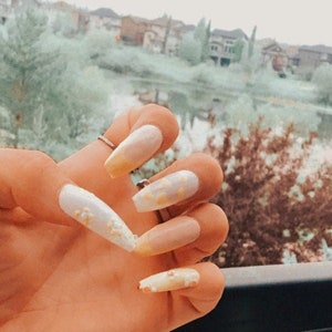 Könnte beinhalten: Nahaufnahme einer Hand mit langen, weißen, mandelförmigen Nägeln, die mit kleinen gelben Blumen verziert sind.