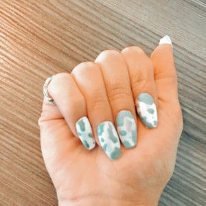 Könnte beinhalten: Nahaufnahme einer Hand mit hellblauem und weißem Kuhflecken-Nagellack auf den Fingernägeln.