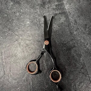 May include: Black and silver hair shears with rose gold finger holes and a pivot screw. The shears are open, displaying the sharp blades. The background is a dark grey, textured surface.