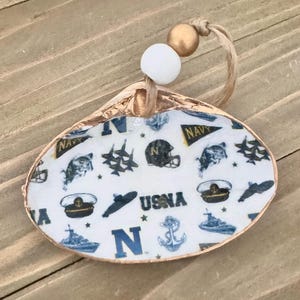 Op de afbeelding: Een schelpenornament met een marinethema. De binnenkant heeft een witte achtergrond met marineblauwe illustraties van schepen, ankers, helmen en de tekst "USNA" en "NAVY". Het heeft een goudkleurige rand, een witte kraal, een gouden kraal en een juten touwtje om op te hangen.