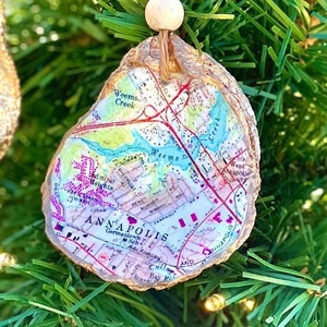 May include: A gold-painted oyster shell ornament with a map of Annapolis, Maryland printed on it. The map is colorful and includes street names and landmarks. The ornament is hanging from a string with white beads.