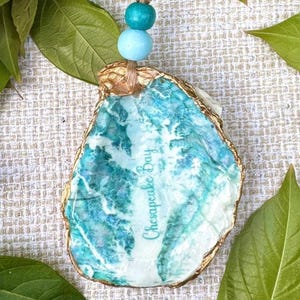 May include: A decorative seashell ornament with a gold trim. The shell has a blue and white marbled pattern and the words "Chesapeake Bay" in teal script. It is suspended from a natural twine cord with blue beads.