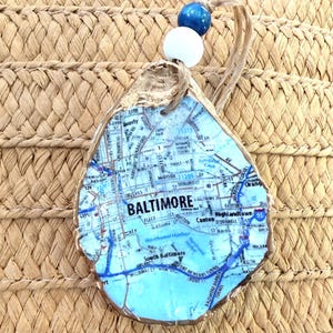 May include: An oyster shell ornament featuring a map of Baltimore, Maryland, with blue and white accents. The ornament is adorned with a blue and white bead and a natural fibre loop for hanging.