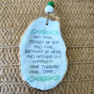 May include: A white, irregular-shaped ornament with gold edging and a jute string. The ornament has the text "May your troubles be less, and your blessings be more, and nothing but happiness come through your door."