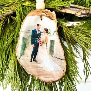 Può includere: Un ornamento rustico in legno con una fotografia stampata di una coppia su un molo. L'ornamento è a forma di pezzo di legno ed è adornato con una corda di perline di legno. Lo sfondo include fogliame verde.