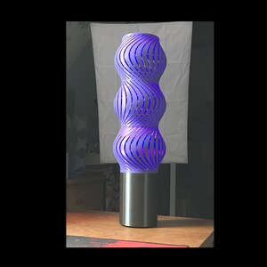 Könnte beinhalten: Eine violette, 3D-gedruckte Lampe mit einem wirbelnden Design. Die Lampe hat einen schwarzen Sockel und strahlt ein violettes Licht aus.