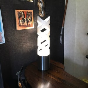 Könnte beinhalten: Eine weiße, moderne Lampe mit geometrischem Design. Die Lampe steht auf einem Holztisch mit Zebra-Muster und einer Schildkrötenfigur an der Wand dahinter.