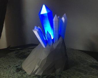 May include: A 3D printed crystal lamp with a blue light emanating from the center. The crystal is surrounded by a gray, rocky base.