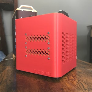 May include: A vibrant red, cube-shaped device with the text "ECI KUBE" on the front. The cube features a honeycomb pattern on the front and side, secured with silver screws. A second, smaller device is visible in the background.
