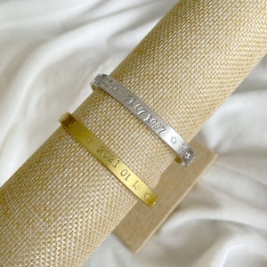 Peut inclure: Deux bracelets en métal gravés, un doré et un argenté, présentés sur une surface neutre. Le bracelet doré porte l'inscription "Joy 2021 01 18" et un symbole de cœur. Le bracelet argenté porte l'inscription "Love 21 07 1997" avec des symboles d'étoiles et de cœurs.