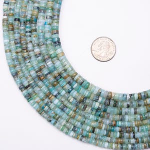 Puede incluir: Primer plano de hebras de cuentas de piedras preciosas azul claro y verde azulado, con una moneda para referencia de tamaño. Las cuentas son pequeñas, cilíndricas y tienen una forma ligeramente irregular. La variación de color incluye tonos de azul, verde y toques de marrón.