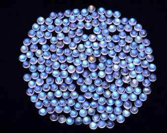 Puede incluir: Una colección de cabochones redondos de piedra lunar iridiscente en varios tonos de azul y blanco. Las piedras están dispuestas en un patrón circular sobre un fondo negro.