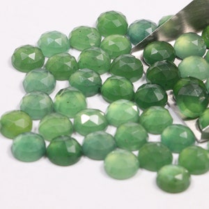 May include: A collection of green gemstone cabochons, with a faceted, rounded shape. The stones are scattered on a white surface, with some spilling from a metal tool.
