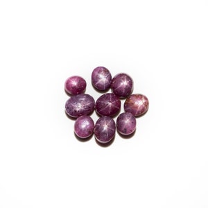 May include: A group of ten star ruby gemstones, each with a six-pointed star, are arranged in a circle on a white background. The rubies are a deep purple-red colour.