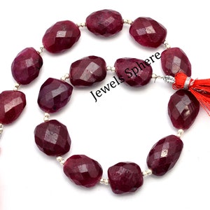 Könnte beinhalten: Eine Kette aus tief roten Rubin-Edelsteinen mit facettierten ovalen Formen. Die Perlen sind auf einem Silberdraht mit silbernen Zwischenstücken aufgefädelt. Die Kette hat an jedem Ende rote Quasten. Das Bild enthält den Text "Jewels Sphere".
