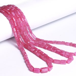 Pode incluir: Um fio de contas de pedras preciosas cor-de-rosa, cada conta é retangular e facetada. As contas são enfiadas em um fio transparente e estão dispostas em um padrão solto e fluido.