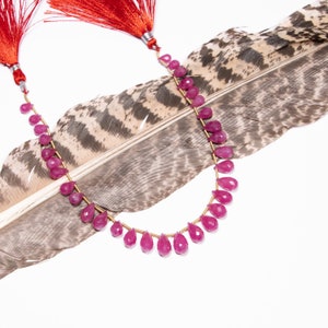 May include: A necklace featuring faceted, teardrop-shaped, pink gemstones strung on a gold-colored wire. Red tassels are at the ends of the necklace. The necklace is displayed on a large, patterned feather against a white background.
