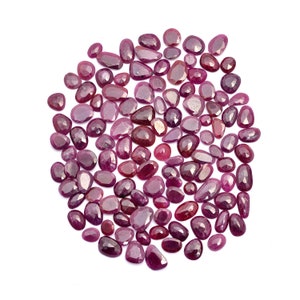 Op de afbeelding: Een grote verzameling losse, gefacetteerde robijn edelstenen in verschillende tinten roze en rood. De stenen zijn verspreid over een witte achtergrond.