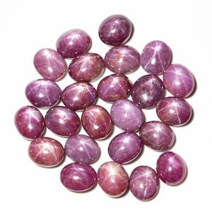 May include: A collection of 20 oval-shaped, pink star ruby gemstones. The stones are arranged in a loose cluster on a white background.
