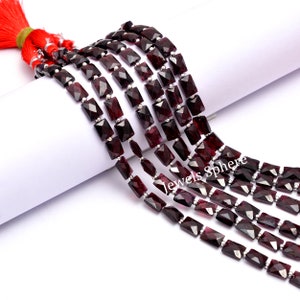 Puede incluir: Una hebra de cuentas de piedras preciosas de color rojo oscuro con forma rectangular facetada. Las cuentas están enhebradas en alambre plateado y tienen aproximadamente 10 mm de longitud. Las cuentas se muestran sobre una superficie blanca.