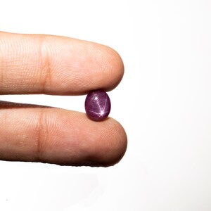 May include: A deep red star ruby gemstone with a six-pointed star in the center. The oval-shaped gemstone is held between two fingers.