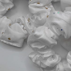 Könnte beinhalten: Eine Sammlung weißer Haargummis mit transparenten Stoffüberzügen. Ein Haargummi zeigt ein Muster aus kleinen weißen Blumen mit goldenen Mitten, während ein anderes mit feinen, schimmernden Glitzerpartikeln im gesamten Stoff versehen ist.