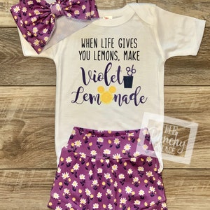 May include: A white baby onesie with the text "When life gives you lemons, make Violet Lemonade" and a purple bow with white flowers. The onesie is paired with purple shorts with white flowers.