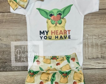 May include: A white baby onesie with a green character holding a red heart and the text "MY HEART YOU HAVE". The onesie is paired with green and yellow shorts featuring a repeating pattern of the same green character holding a red heart.