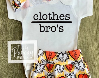 May include: A white baby onesie with the text "clothes bro's" printed on it. The onesie is paired with a headband and shorts with a red, yellow, and white heart pattern.