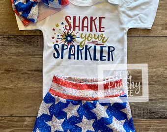 May include: A white baby bodysuit with the text "Shake Your Sparkler" in red, gold, and blue. The outfit includes blue star-patterned shorts and a matching bow headband, all in patriotic colors.