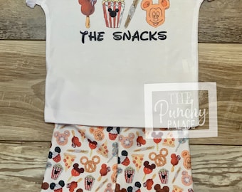 May include: White t-shirt with the text "I'm here for the snacks" and a Mickey Mouse head with various snacks. Matching white shorts with a Mickey Mouse head and snack print.