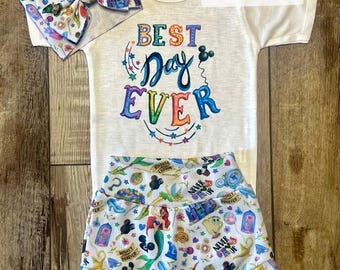 May include: A white baby onesie with the text "Best Day Ever" in rainbow colors. The onesie is paired with white shorts featuring a colorful print of Disney characters and icons. A white headband with a colorful Disney print is also included.