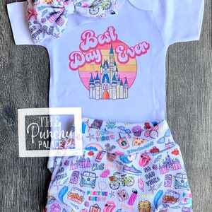 May include: A white baby outfit set with a matching headband. The onesie features the text "Best Day Ever" with a castle graphic. The shorts and bow have a colorful pattern with various icons.