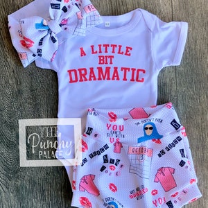 May include: A white baby onesie with the text "A Little Bit Dramatic" in red. A white headband with pink lips and a calendar print. White shorts with a pink and blue print featuring the text "You Can't Sit With Us" and "So Fetch".
