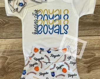 May include: A white baby onesie with the text "Kansas City Royals" in blue and gold. A white headband with a baseball and the word "Royals" in blue and gold. White baby shorts with a baseball and the word "Royals" in blue and gold.