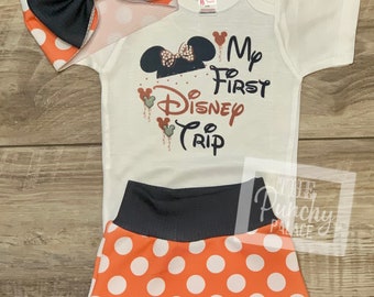 May include: A white baby onesie with black and orange polka dots and the text "My First Disney Trip" with a Mickey Mouse head graphic. The onesie is paired with a black and orange polka dot headband and orange and black polka dot shorts.