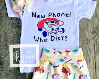 May include: A white baby onesie with the text "New Phone! Who Dis??" and a cartoon image of a red toy phone. The onesie is paired with a pair of yellow shorts with a pattern of toy phones and other toys.
