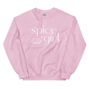 May include: A light pink sweatshirt with the words "spice girl" in white script. A white illustration of an open book with sparkles is below the words. The sweatshirt is a crew neck style.