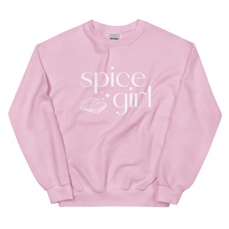 May include: A light pink sweatshirt with the words "spice girl" in white script. A white illustration of an open book with sparkles is below the words. The sweatshirt is a crew neck style.