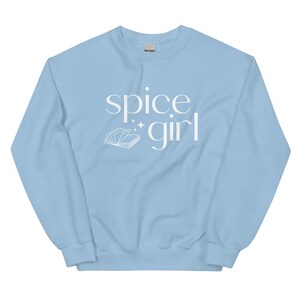 May include: A light blue sweatshirt with the words "spice girl" in white script. A white illustration of an open book with sparkles is next to the word "girl". The sweatshirt is made of a soft material.