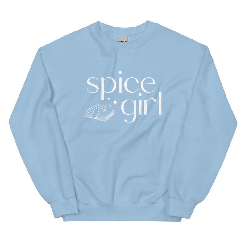 May include: A light blue sweatshirt with the words "spice girl" in white script. A white illustration of an open book with sparkles is next to the word "girl". The sweatshirt is made of a soft material.