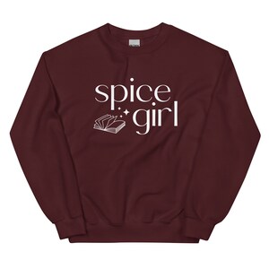 May include: A maroon sweatshirt with the words "spice girl" in white script. A small open book graphic and stars are next to the word "girl". The sweatshirt is a solid color and has a crew neck.