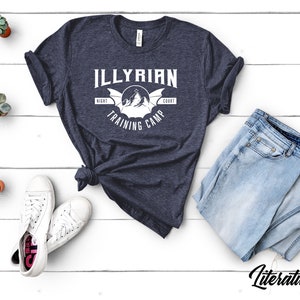 Puede incluir: Una camiseta azul marino con un diseño gráfico blanco que dice "Illyrian Night Court Training Camp". El gráfico presenta un murciélago estilizado con montañas en el fondo.